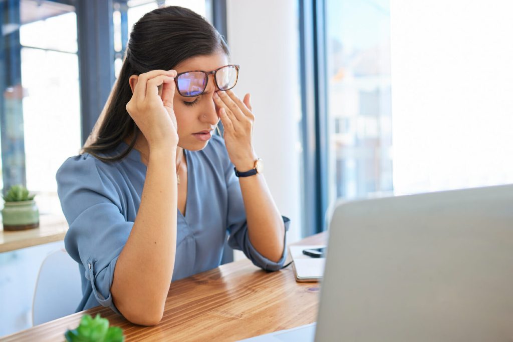 Woman experiencing discomfort while wearing glasses, rubbing her temples at a desk with a laptop, indicating potential headache issues related to eyewear.